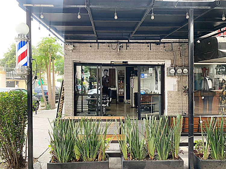 Entrada de Mr. Gaboss bajo el portal negro con focos colgantes, poste de barbero rojo, blanco y azul y jardineras al frente